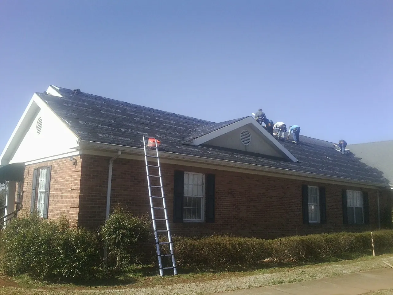 Crew tearing shingles off a brick ranch roof with a ladder set — Emergency Roof Repair work by our crew in South Carver