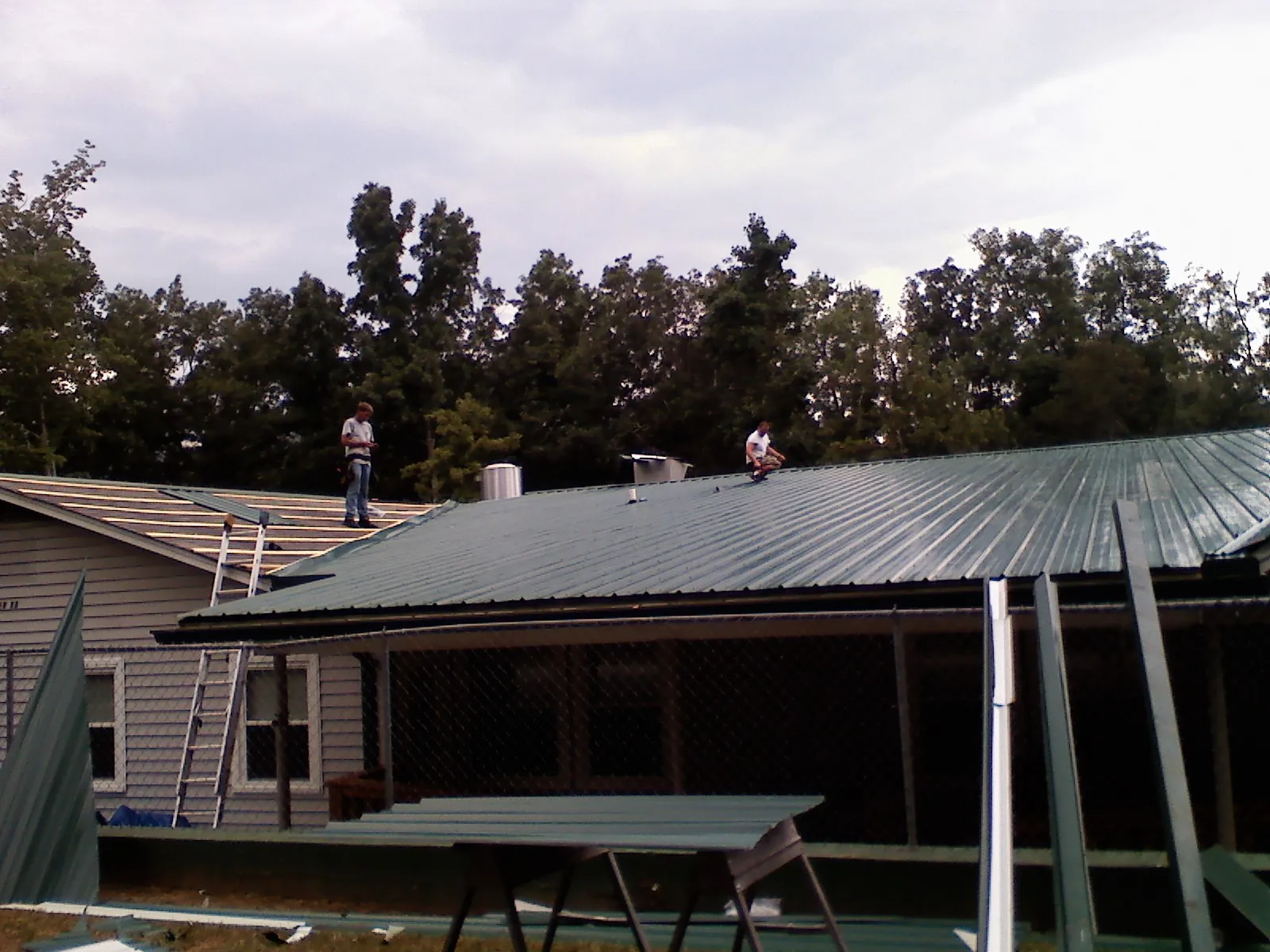 Two roofers installing green metal panels above a covered porch in South Carver